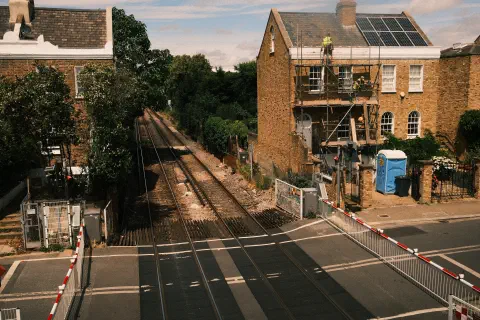 A photo looking down on a level crossing. The barriers are closed. On one side, workmen work on the roof of a house.
