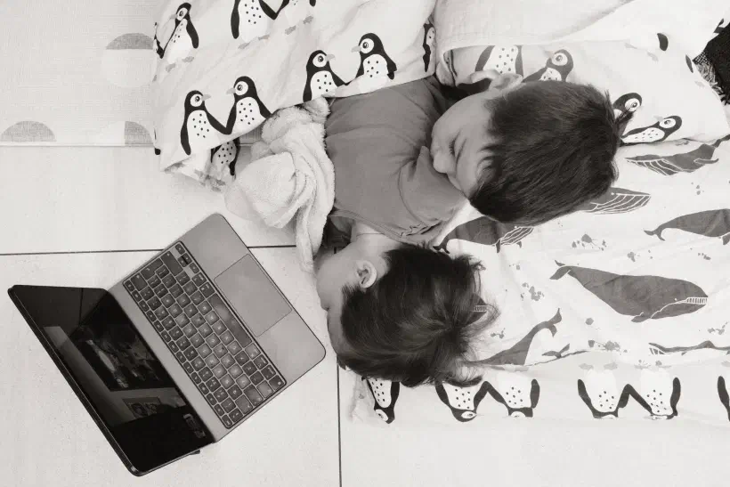 A black and white photo of two boys laying on the ground under a duvet looking at an iPad.
