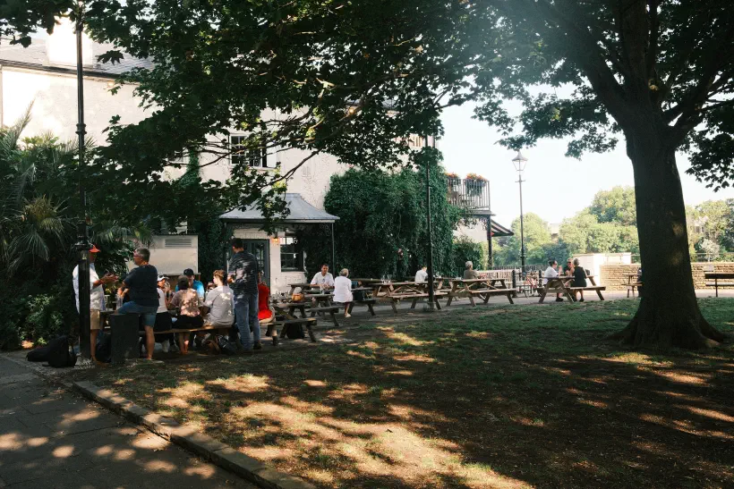 A shaded area outside a pub. In corner, group of people sit on a pub table.