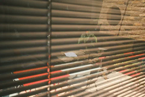 A photo of a partial reflection in a window. The gaps between the blinds allow light from outside to shine through, but the reflection of a table inside the room can also be seen in the glass.