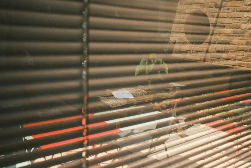 A photo of a partial reflection in a window. The gaps between the blinds allow light from outside to shine through, but the reflection of a table inside the room can also be seen in the glass.