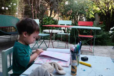 A boy sits at a small table, in front of him a colouring book and a pot of pens, a garden table and chairs can be seen in the background