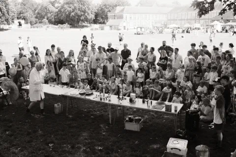Black and white and white photo of a crowd of people facing towards a row of tables. On the tables, an array of scientific apparatus. In front of them, an old man in a white lab coat performs experiments.