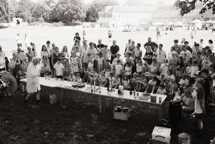 Black and white and white photo of a crowd of people facing towards a row of tables. On the tables, an array of scientific apparatus. In front of them, an old man in a white lab coat performs experiments.