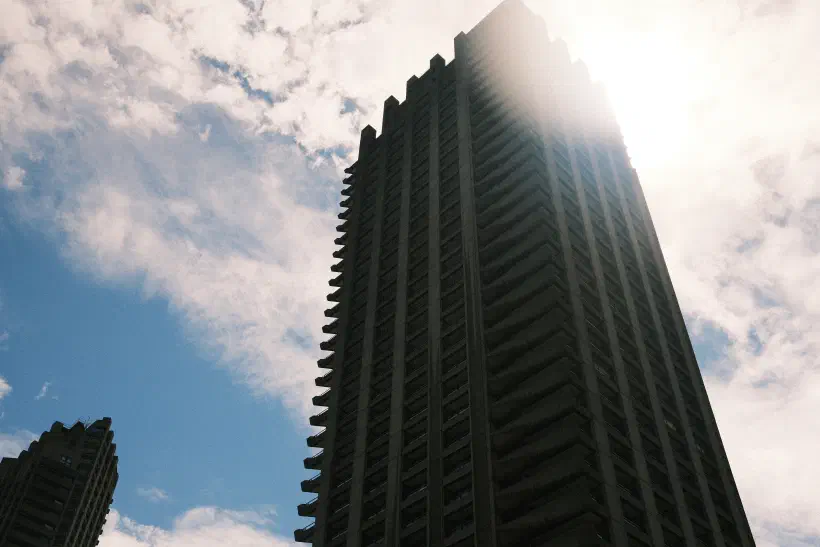 A photo looking up at the midday sky. A tall skyscraper in the foreground. The bright sunlight blooms over the edge of the building.