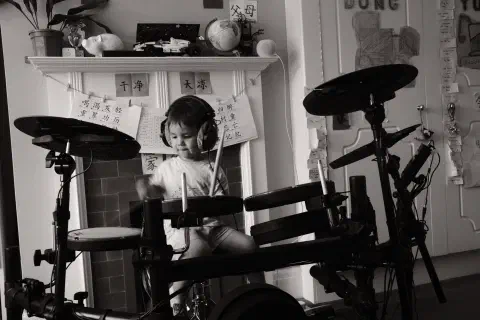 A black and white photo of a young boy playing an electronic drum kit. On the wall behind him, Chinese characters hang from a piece of string.