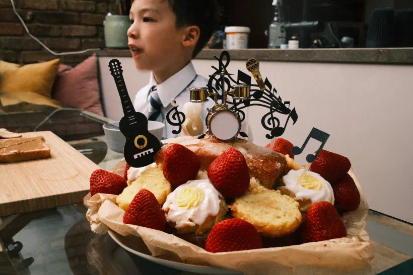 A photo of a birthday cake. The cake is sponge with lemon icing. It is decorated with strawberries, musical notes, a guitar and a drum kit. The candle indicates it is an eighth birtday cake. Behind it a boy looks out of the frame. We don't see what he is looking at.
