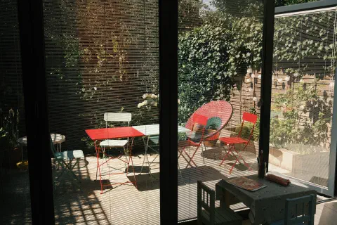 A photo looking out through glass doors to a garden. Despite the blinds being closed, the bright sun outside lights the garden leaving the room in the dark.