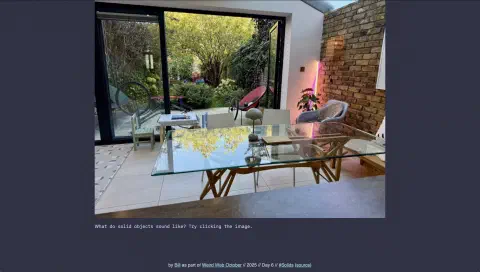 A photo of a dining room that opens out onto a garden. The table in the centre of the room is glass with a wooden frame. The doors to the garden are open. In the far corner, two vertical strip lights are on. The right hand wall of the room is bare brick, the far wall is painted white.