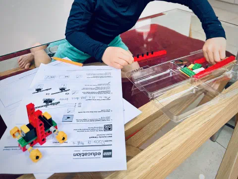 A child sits at a glass table. In front of them a tray of lego pieces and a printed worksheet. On top of the worksheet is a small wheeled catapault. The main beam is not yet attached. The child is adding pegs to join two halves of the beam together.