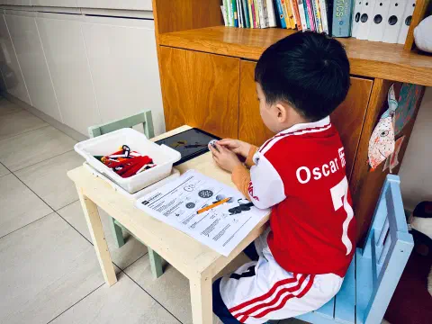 A boy in a red football shirt sits at a small table next to a bookshelf. On the table in front of him is a tray of Lego, a worksheet and an iPad showing LEGO instructions. He is holding a LEGO gear in his hands.